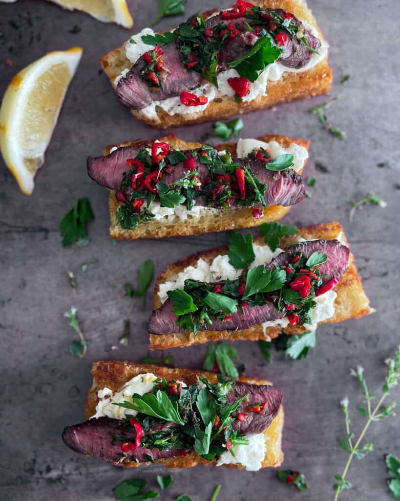 Sliced bread, toasted and topped with burrata cheese, steak, peppers and parsley. Dark sheet pan background with oregano flowers.