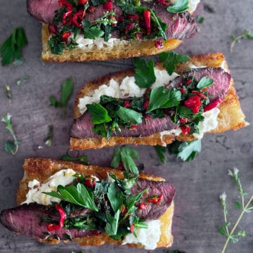 Sliced bread, toasted and topped with burrata cheese, steak, peppers and parsley. Dark sheet pan background with oregano flowers.