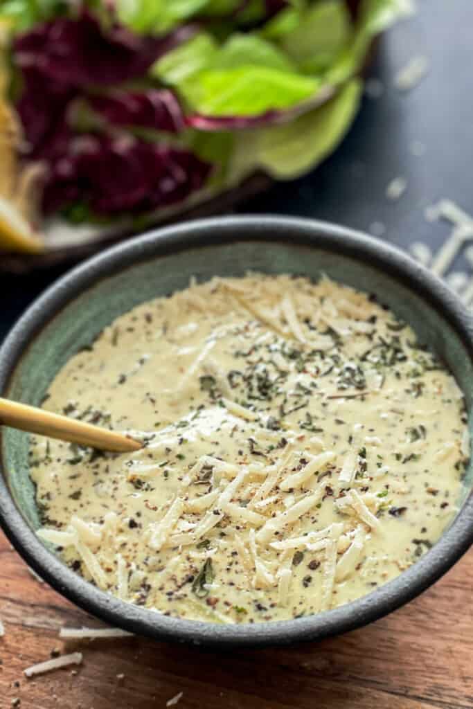 Caesar dressing in a bowl topped with parmesan cheese and dried herbs with salad green in the top left corner.
