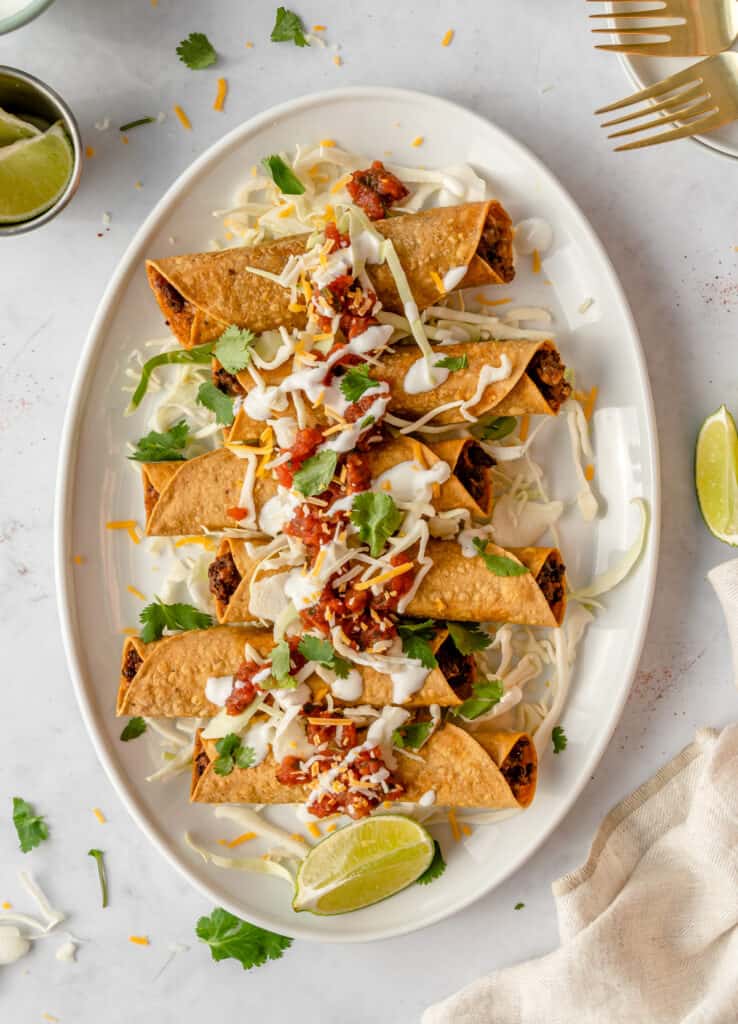 Beef taquitos on a white oval plate served with limes, herbs and sour cream