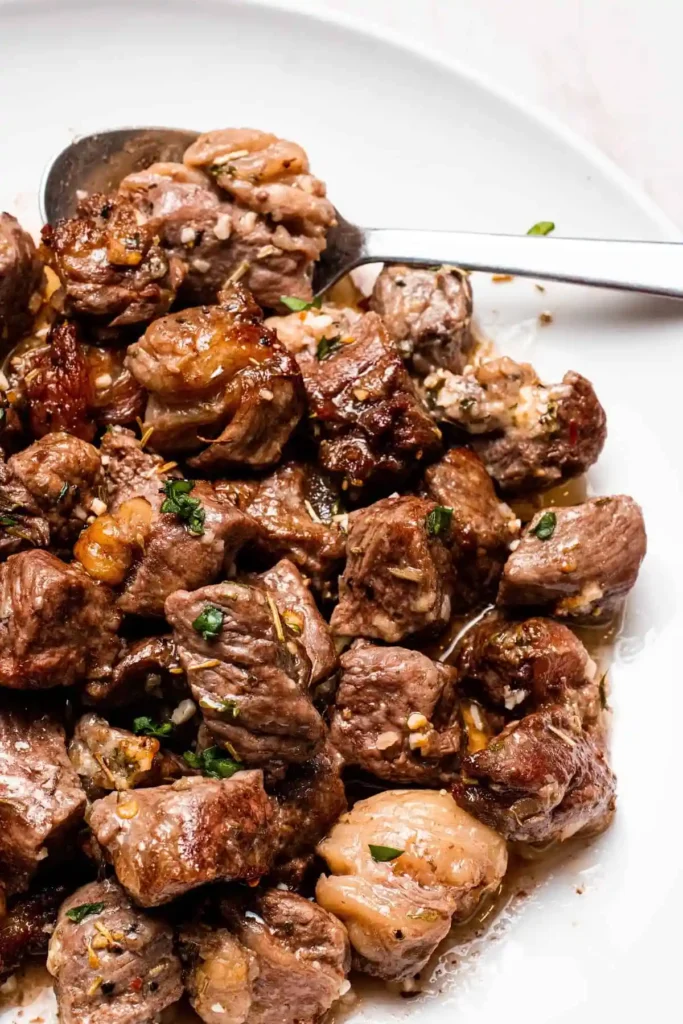 Steak bites covered in butter and herbs on a white plate with a spoon and a white background