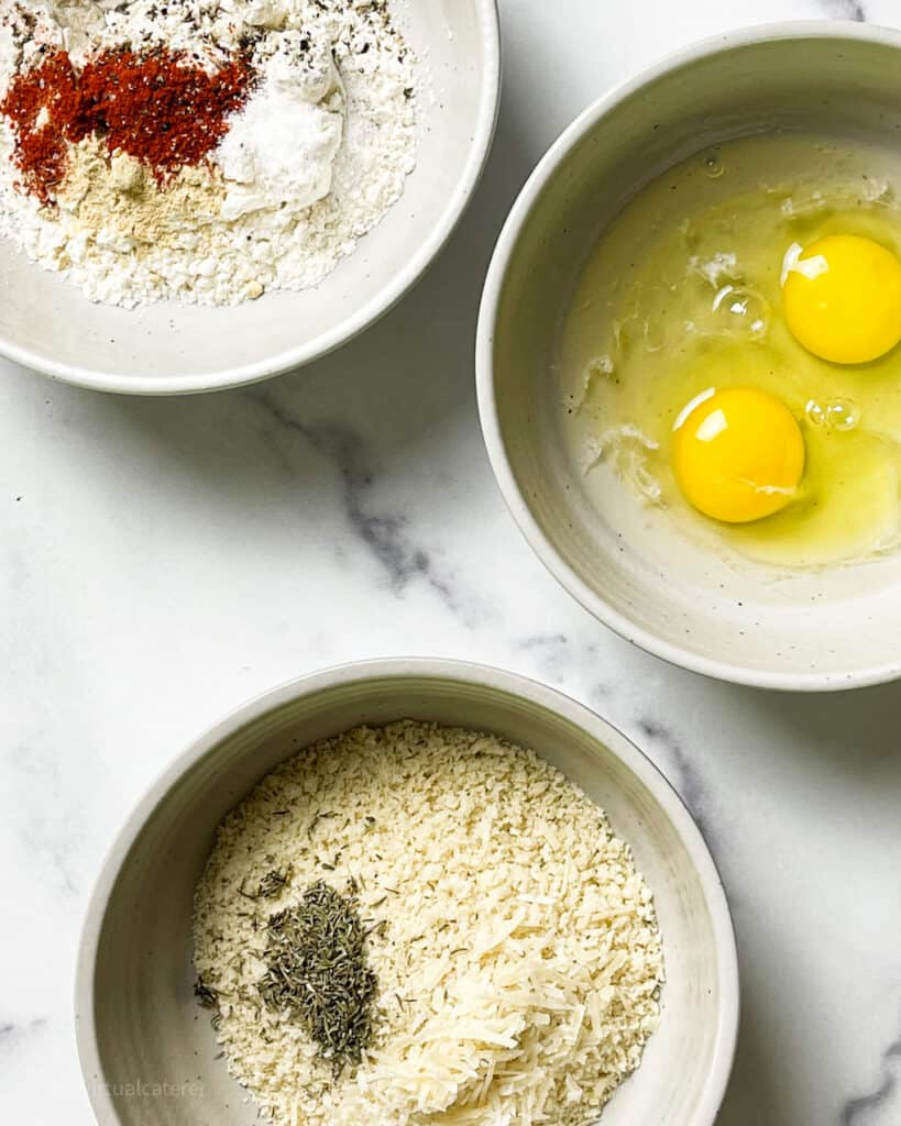Breading station.  3 bowls on white marble background.  One bowl with flour and spices, anther with eggs and the last with breadcrumbs and Italian seasoning
