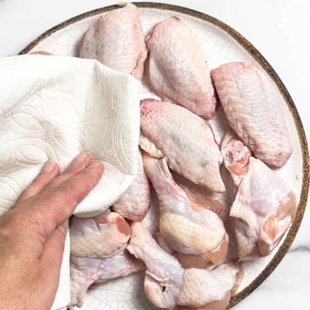 patting raw chicken wings dry on a white plate with a brown rim on a white background