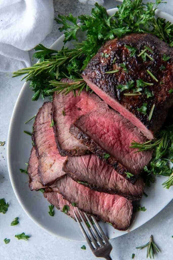 Roast beef sliced on a white plate with rosemary and parsley garnishes