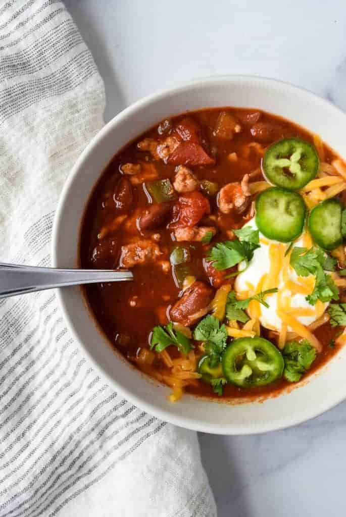 Chili in a bowl with a spoon topped with jalapenos, cheese and sour cream.