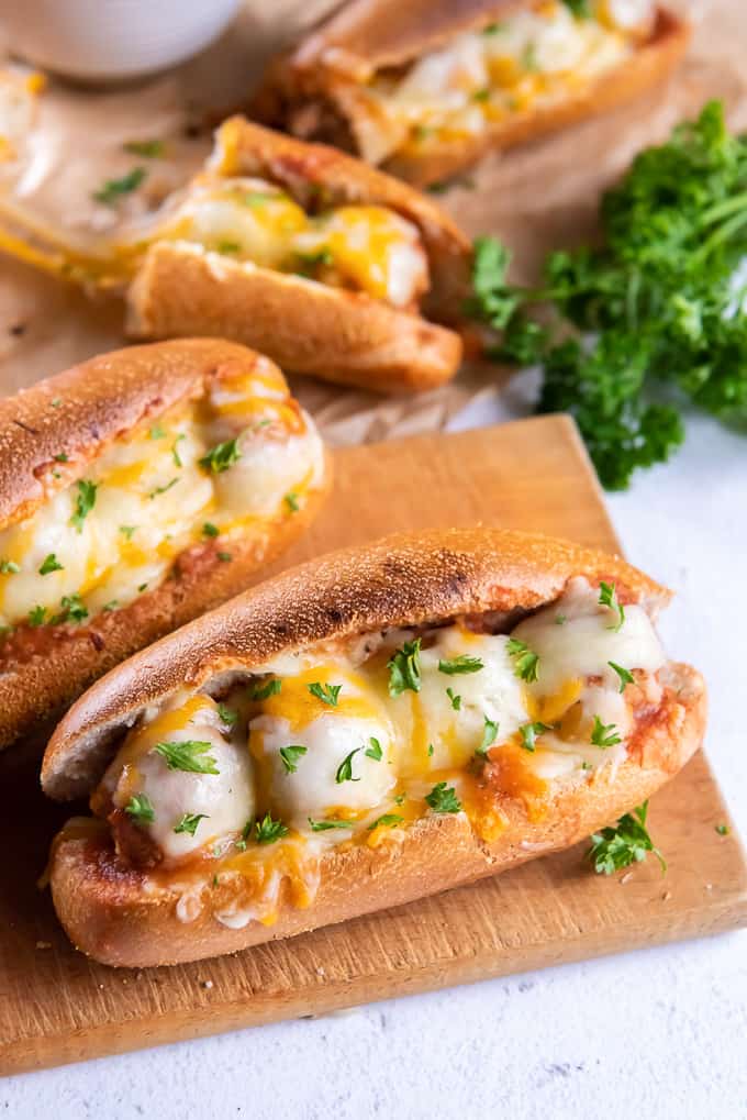 Meatballs subs sitting on a cutting board on a white counter.  Melted cheese covers the meatballs between 2 crusty pieces of bread.