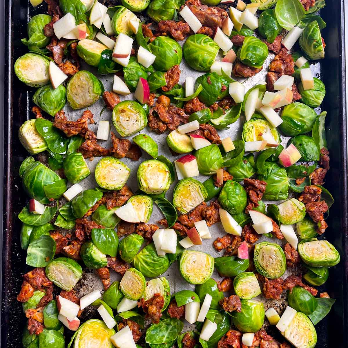 Brussels sprouts with sausage, apples and seasonings on a sheet tray getting ready to roast.