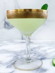 A side angle shot of a cucumber martini in a martini glass with a gold rim garnished with a mint leaf and cucumber slice.