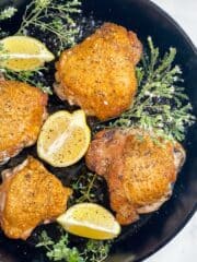 A cast iron skillet filled with cooked crispy chicken thighs and garnished with lemon wedges and bunches of fresh herbs.