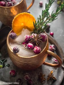 A cocktail filled with ice in a copper mug garnished with sugared cranberries, orange slices, fresh rosemary, and star anise. Dark grey background with sugared cranberries.