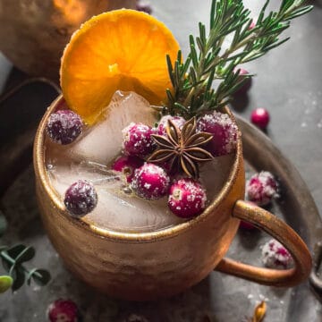 A cocktail filled with ice in a copper mug garnished with sugared cranberries, orange slices, fresh rosemary, and star anise. Dark grey background with sugared cranberries.