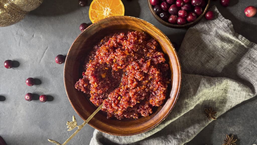A wooden bowl filled with cranberry relish is placed on a gray cloth. A spoon rests in the bowl. Nearby are fresh cranberries, half a cut orange, and star anise on a dark surface.