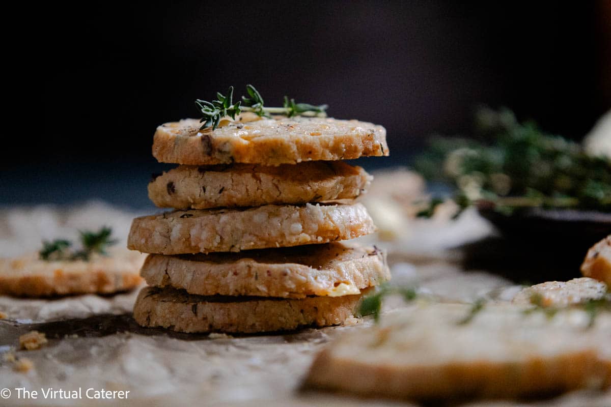A stack of savory cookies on parchment paper with fresh thyme and cheddar cheese.