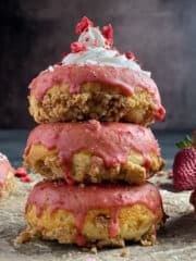 A stack of donuts on with pink icing running down the sides on parchment paper. Topped with cream cheese frosting and dried strawberries from the side view.