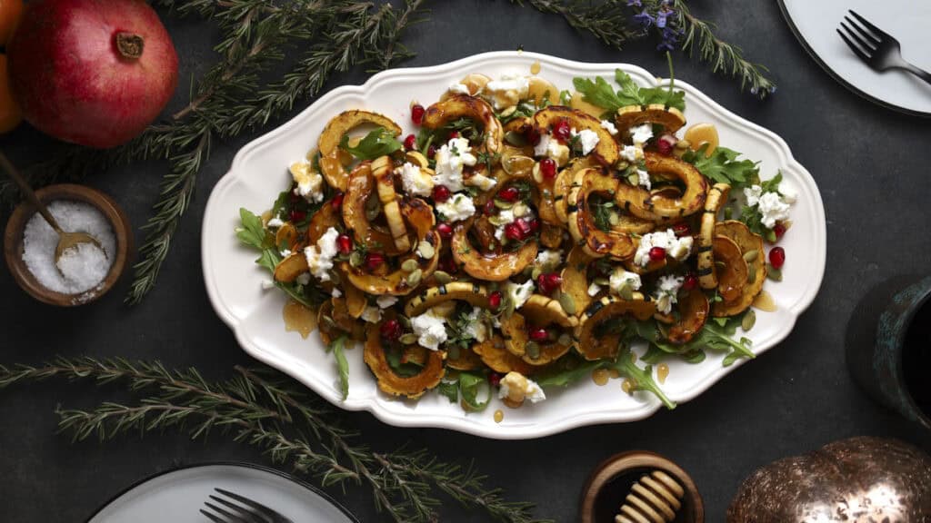 A festive salad on a white platter, featuring roasted squash slices, goat cheese crumbles, pomegranate seeds, and greens, garnished with rosemary sprigs. Flanked by a pomegranate, salt dish, and honey dipper on a dark table.