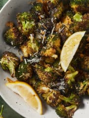 Parmesan roasted broccoli in a bowl served with lemon wedges on a green background.