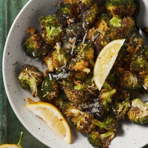 Parmesan roasted broccoli in a bowl served with lemon wedges on a green background.