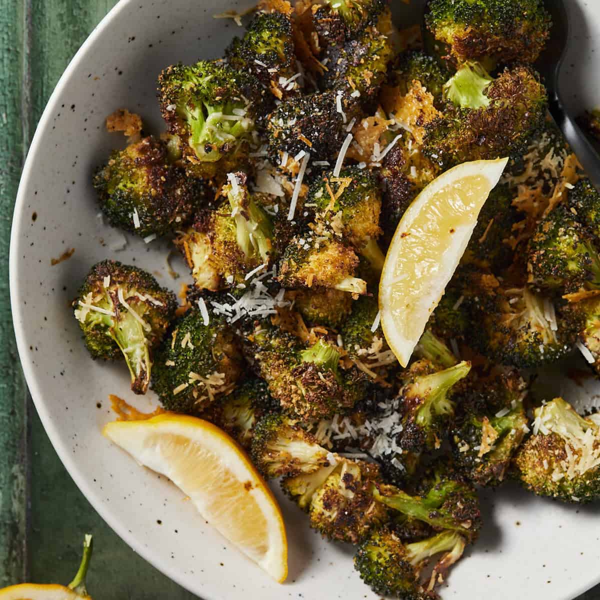 Parmesan roasted broccoli in a bowl served with lemon wedges on a green background.