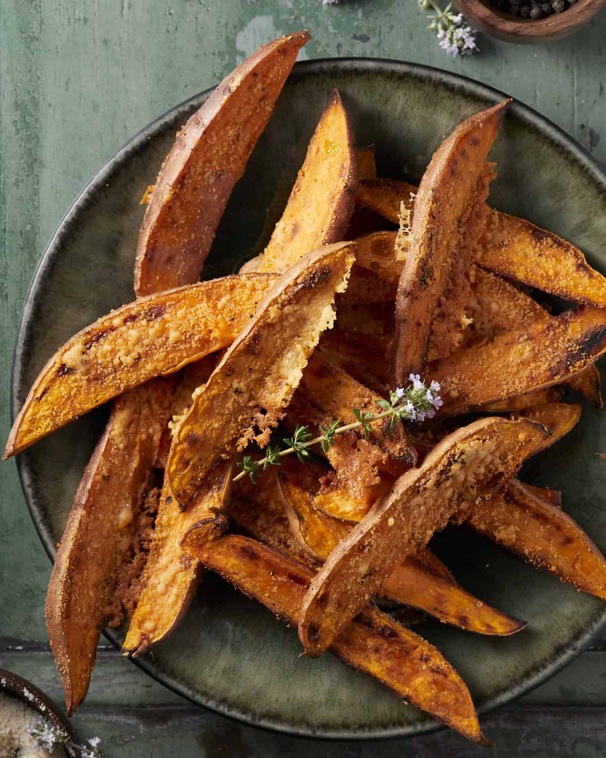 Sweet potato wedges crusted in parmesan cheese on a plate with fresh thyme. 