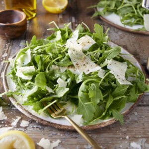 A simple arugula salad with strip of parmesan cheese on a rustic plate and side of lemon.