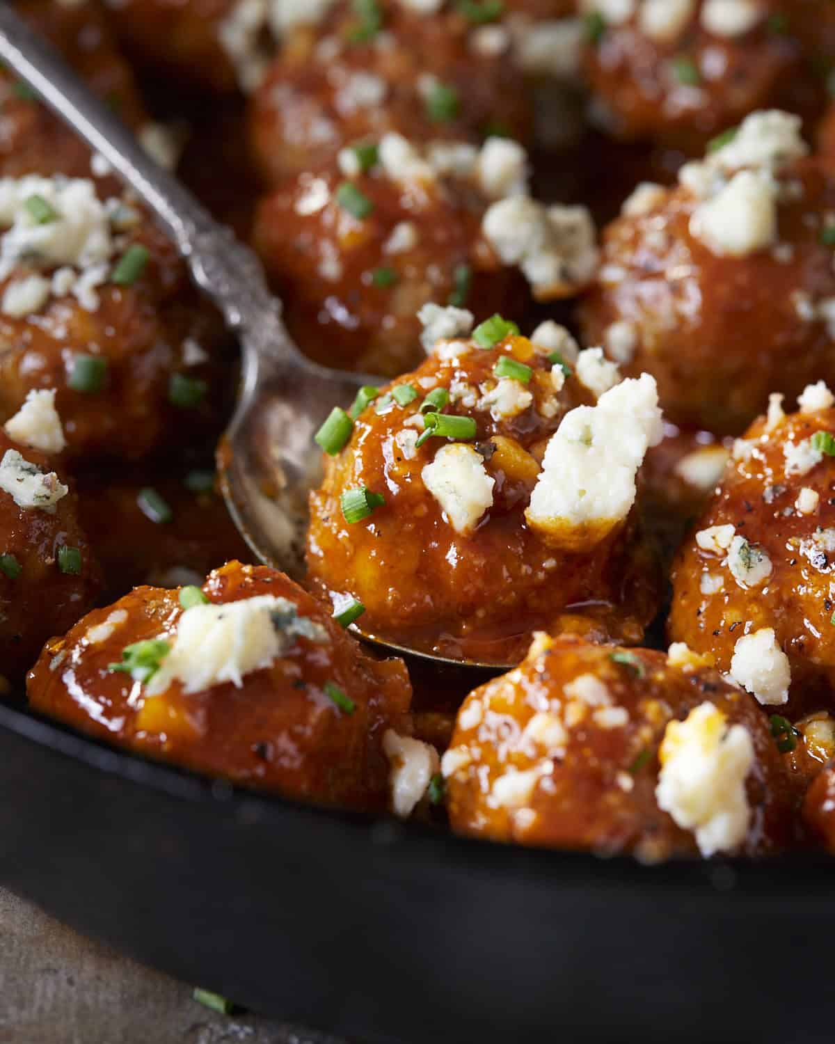 Up close of a buffalo chicken meatball topped with blue cheese and chives in a spoon.