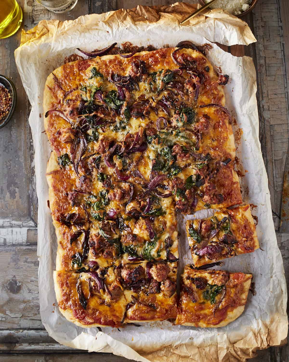 A baked sheet pan pizza with rustic parchment paper sliced with sausage, onions and chard. 