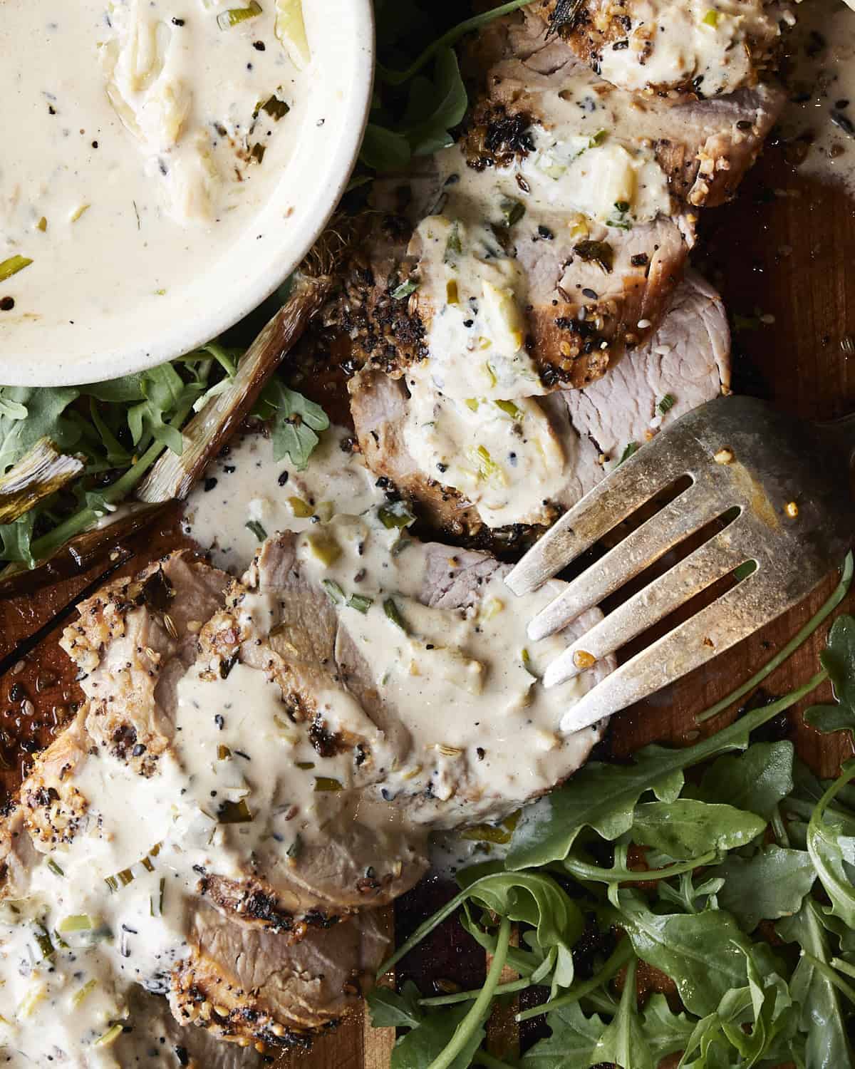 Up close of sliced pork tenderloin with a fork and a side of sauce and arugula.