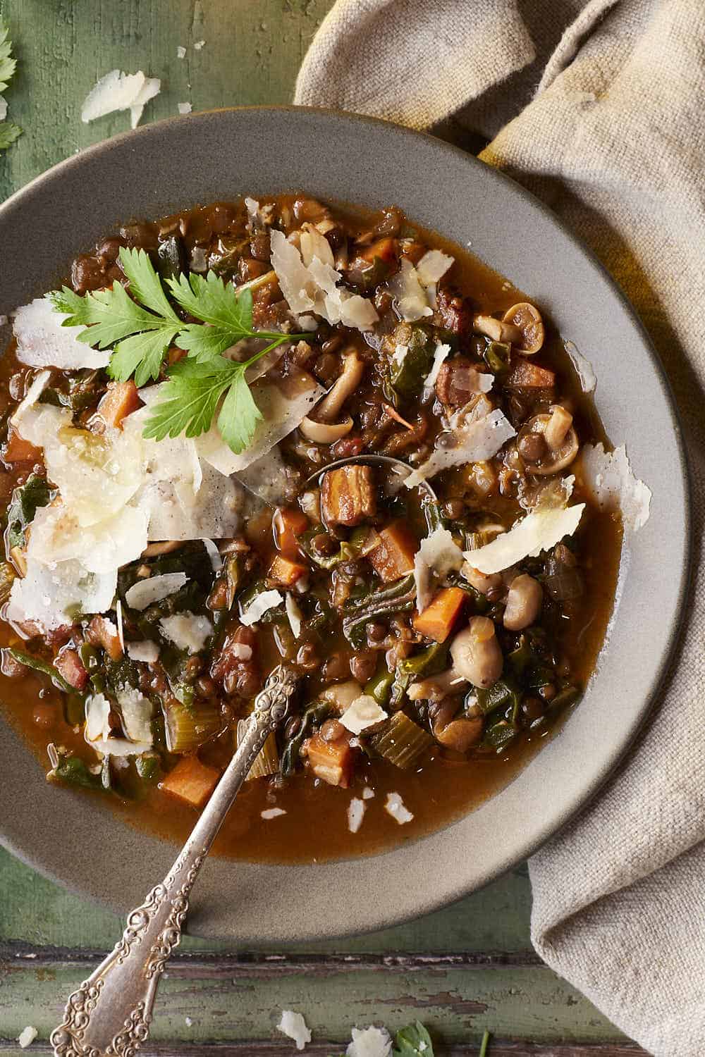 A bowl filled with a bacon lentil soup with lots of veggies and a spoon.  It's topped with parmesan and parsley. 