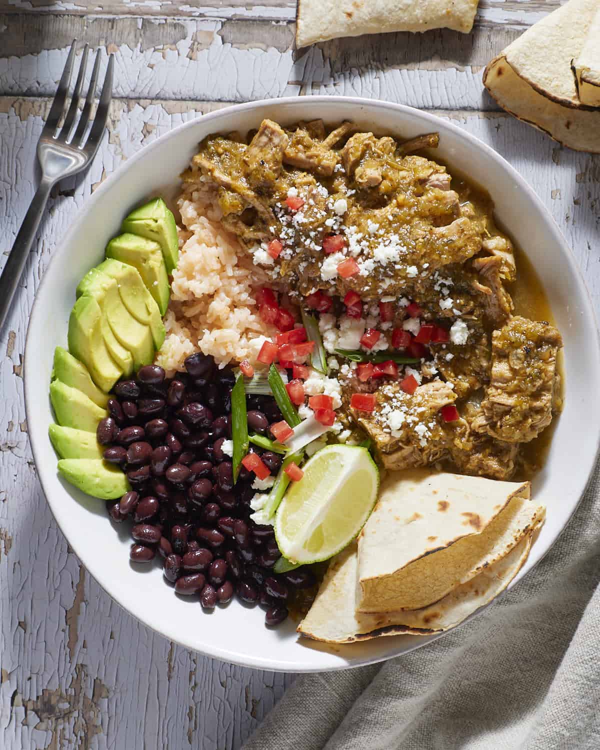 A white bowl filled with chile verde pork, black beans , rice and avocado.