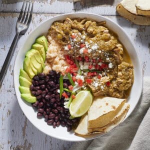 Chile verde in a bowl with beans, rice, avocado and garnishes.